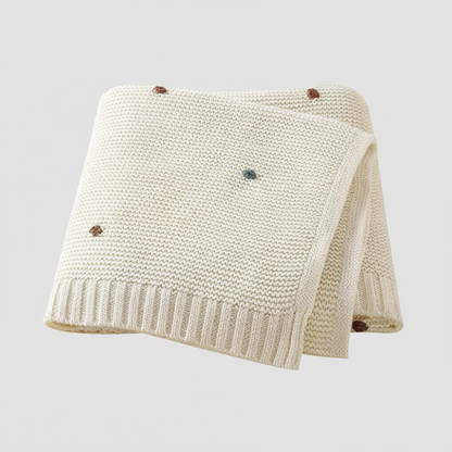 Couverture bebe tricot crème points brodés colorés pliée sur fond gris clair