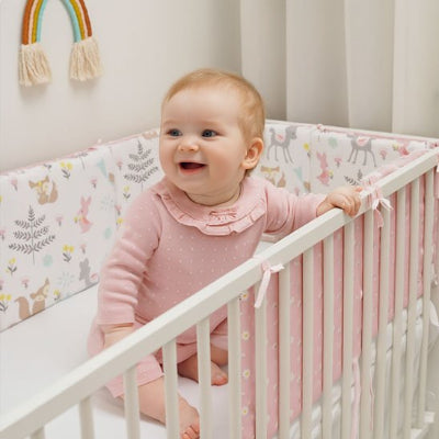 Bébé souriant assis dans lit blanc avec tour de lit  rose motifs animaux safari pastel, chambre décoration arc-en-ciel macramé.